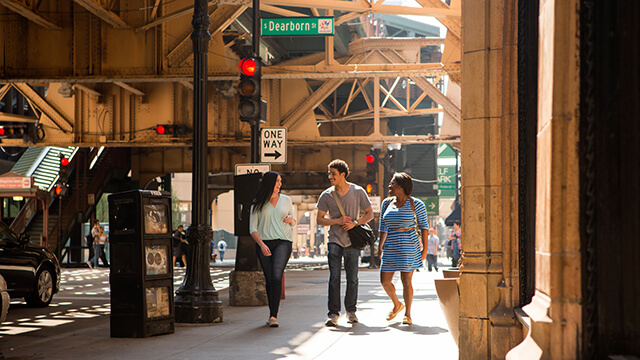 three people walk on a sidewalk underneath an elevated train track