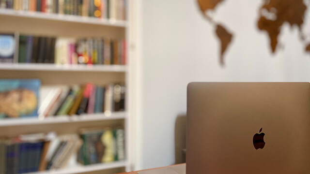 an open laptop sits on a desk in front of a bookshelf and a map of the world mounted on the wall