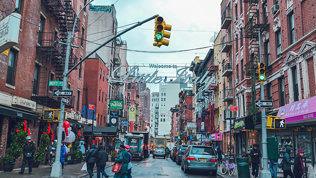 a sign reading "welcome to little italy" hangs over a street lined with cars and brick buildings