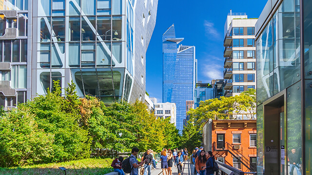 people walking along the highline, an elevated park, in New York City. Skyscrapers tower on either side of the park, which is covered in greenery.