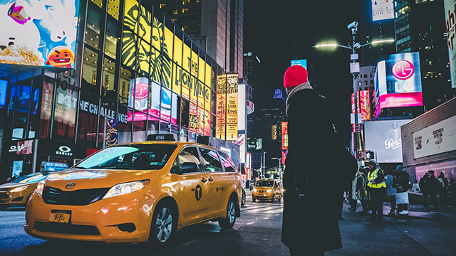 a man in a coat and red hat stands on the sidewalk in front of brightly lit billboards and a yellow taxi