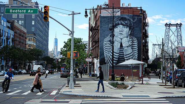 people cross a street in front of a tall brick building with a black-and-white mural painted on its side and a bridge in the background