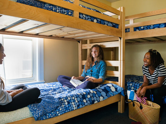 three strangers sharing a hostel dorm room sit talking on wooden bunk beds with blue bed spreads
