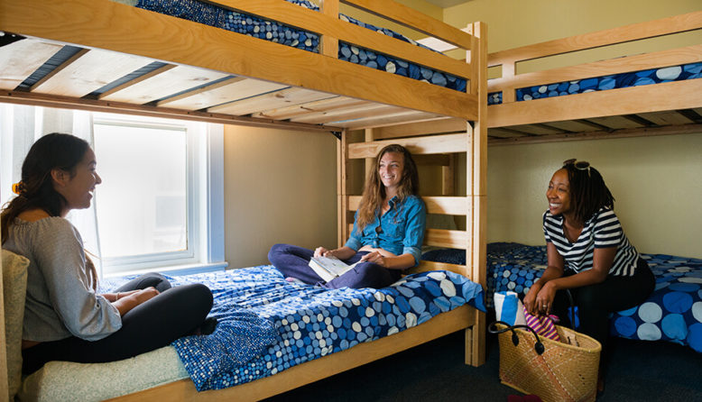 three strangers sharing a hostel dorm room sit talking on wooden bunk beds with blue bed spreads