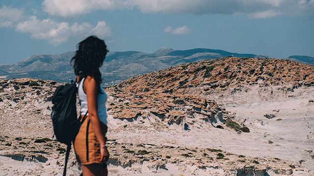 a side view of a young woman in a white tank top and tan shorts wearing a small black backpack. She is looking out over sand and rocks with brown and green hills in the background.