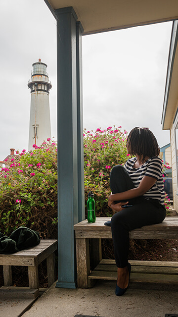 a guest at HI Pigeon Point Lighthouse hostel sits looking out over the  historic pigeon point lighthouse. She is wearing black pants and a black and white striped shirt. 