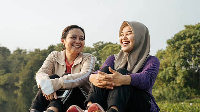 two confident female travelers sit side by side laughing together. Behind them are green trees. 