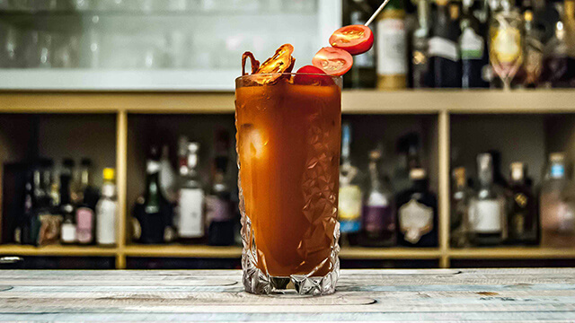 A reddish-brown-colored cocktail in a tall glass garnished with cherry tomatoes sits on a marble bar
