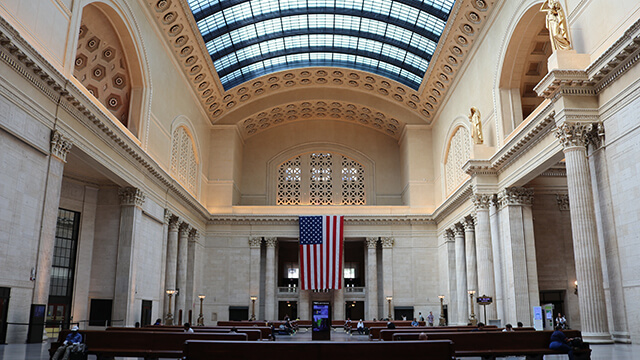 a large white hall with a domed glass ceiling and a hanging american flag inside Chicago's union station. 