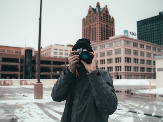 a man in a winter coat and hat takes a photo with a large camera while standing in front of brick buildings on a day trip from chicago to milwaukee