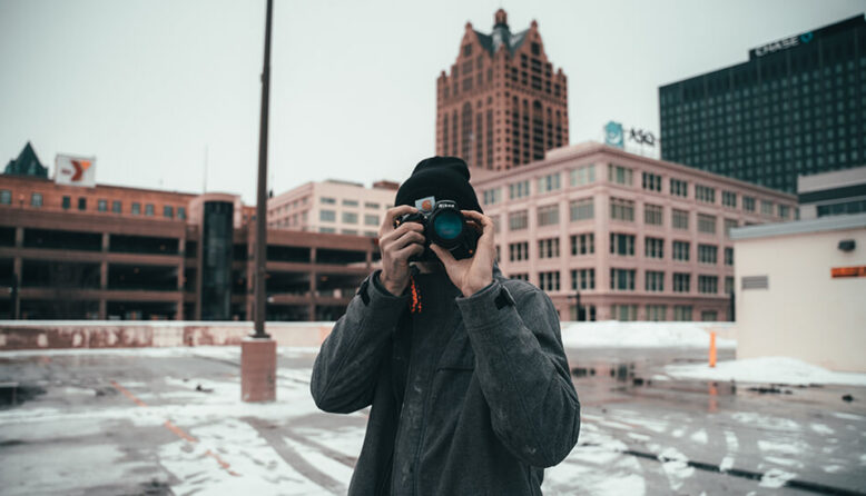 a man in a winter coat and hat takes a photo with a large camera while standing in front of brick buildings on a day trip from chicago to milwaukee