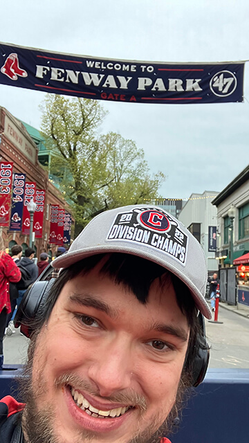 cleveland guardians fan Peter Knab in boston poses beneath an outdoor banner for Fenway Park