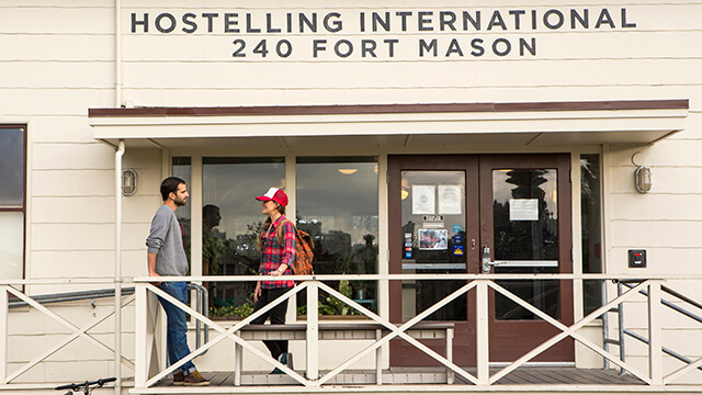 a man and a woman stand talking in front of the door to a historic white building that houses HI San Francisco Fisherman's Wharf hostel