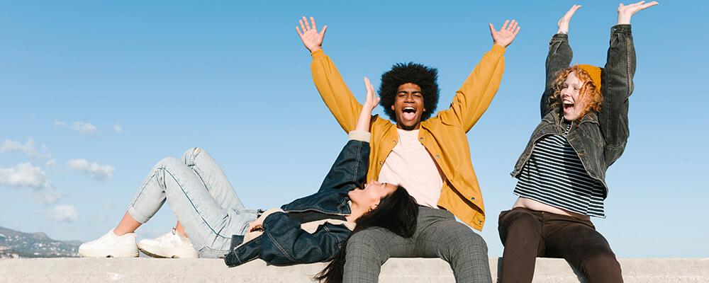 three young friends outside with their hands in the air and a blue sky