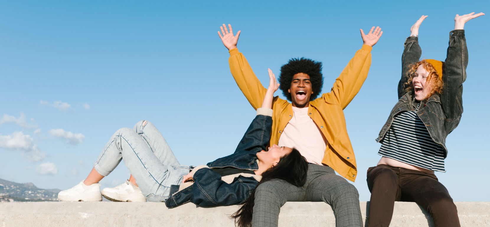 three young friends sit on a cement wall with their arms in the air