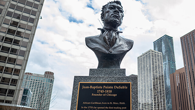 a bust of Jean Baptiste Pointe DuSable, the founder of Chicago, overlooking the Chicago River. Skyscrapers and a blue sky with white clouds are in the background.