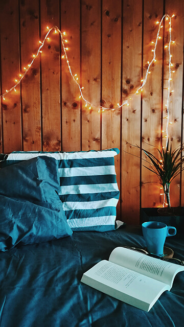 a bed with blue pillows against a wood-paneled wall with string lights hanging above
