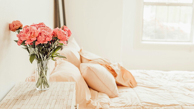 a bouquet of pink flowers on a bedside table beside a bed with cream-colored sheets and pillows