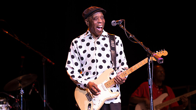 the legendary guitarist Buddy Guy plays a cream-colored guitar and sings into a microphone. He is wearing a white shirt with black polka dots.