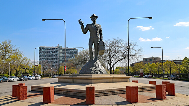 the monument to the great northern migration in Chicago. A statue of a man with one had raised in the air and his other hand carrying a suitcase, surrounded by a ring of suitcases, against a blue sky.
