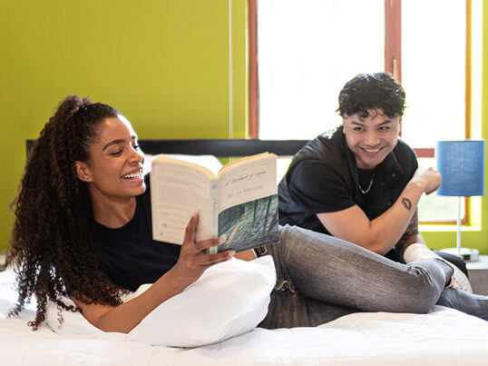 a happy couple lays on a queen-sized bed in a private room at a a HI USA hostel laughing and looking at a book