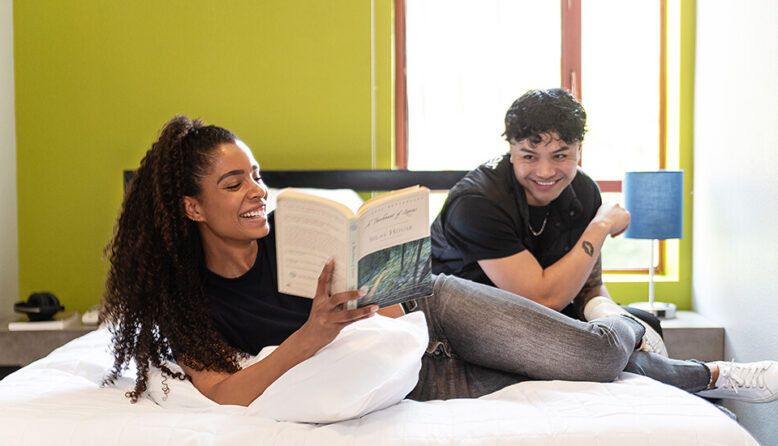 a happy couple lays on a queen-sized bed in a private room at a a HI USA hostel laughing and looking at a book