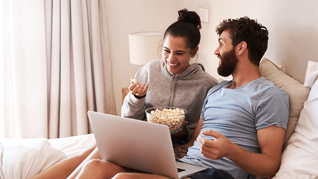 a young couple sitting in bed watching a movie on a laptop while eating popcorn