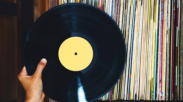 a hand holds up a single vinyl record with a yellow label. On the shelf behind are the edges of dozens of colorful record covers.