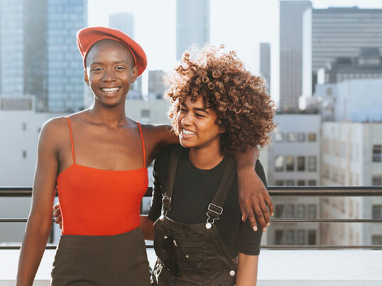 two young friends laughing and smiling with their arms around each other standing on a rooftop overlooking the city skyline