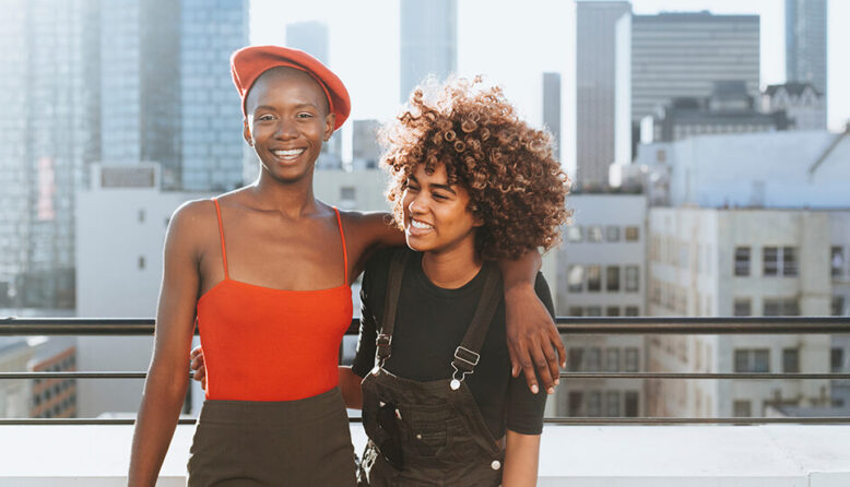 two young friends laughing and smiling with their arms around each other standing on a rooftop overlooking the city skyline