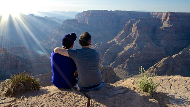 two young men sit looking out over the Grand Canyon. One has his arm around the other's shoulders.