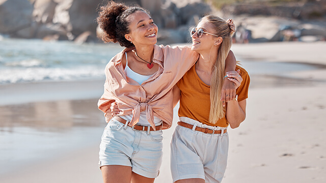 two young women walking on the beach with their arms around each other's shoulders