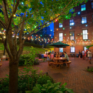 HI New York City hostel's private courtyard at dusk. Groups of travelers sit at round picnic tables, with multi-colored lights hanging from the green trees.