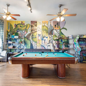 A pool table at HI San Diego Downtown hostel sits atop a wood floor with a vibrant multi0colored mural behind it and arcade games to the right.
