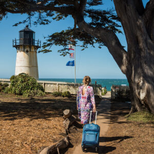 A guest pulling a small suitcase behind her walks down a path at HI Point Montara Lighthouse hostel, with the ocean, blue sky, and Point Montara lighthouse ahead of her.