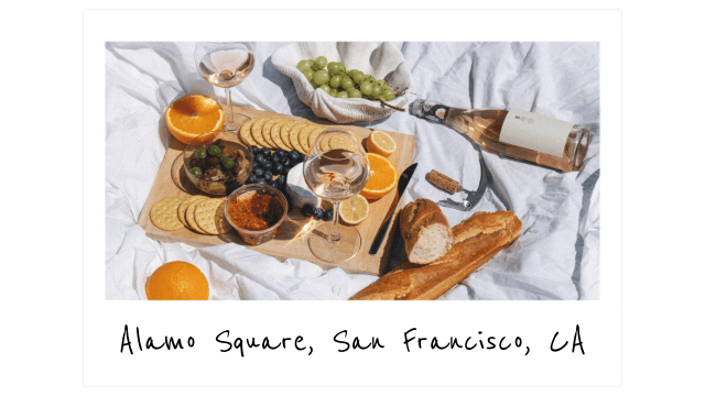 a white picnic blanket with glasses of rose wine, a baguette, and a cutting board covered in olives and crackers