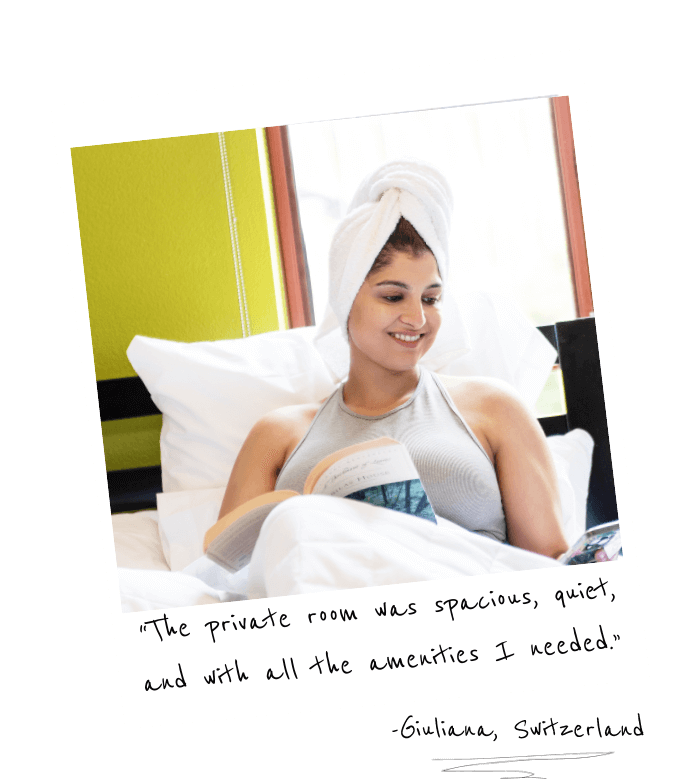 a polaroid photo of a young woman in a large bed with crisp white sheets. She has a towel wrapped around her wet hair and is smiling while looking at her phone. There is an open book on her lap. The writing on the photo reads,