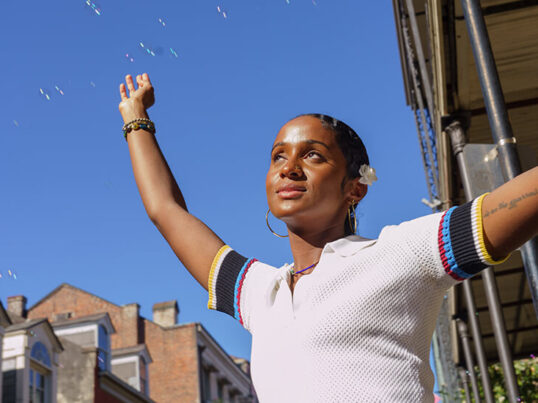 a young woman stands in the sunshine with her arms spread out as bubbles float through a blue sky