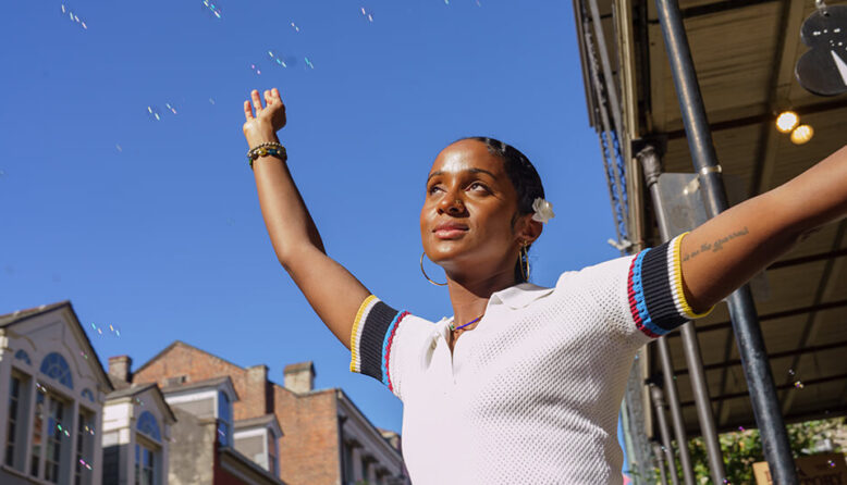 a young woman stands in the sunshine with her arms spread out as bubbles float through a blue sky