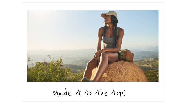 a woman in a baseball hat, tank top, and shorts sits on a boulder at the top of a mountain in San Diego