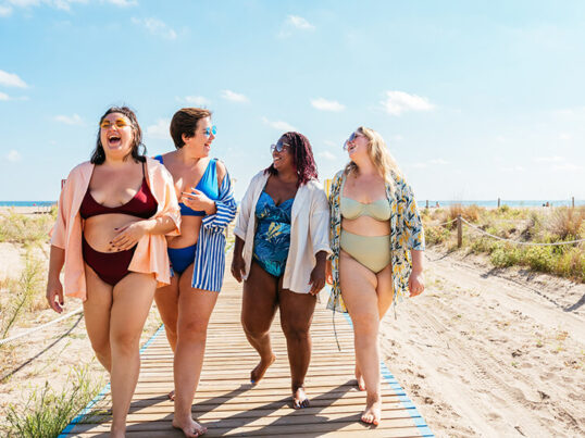 four women in swimsuits laugh as they walk down a boardwalk on the beach on Cape Cod