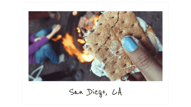 a polaroid of a hand with light blue nail polish holding a s'more in front of a bonfire