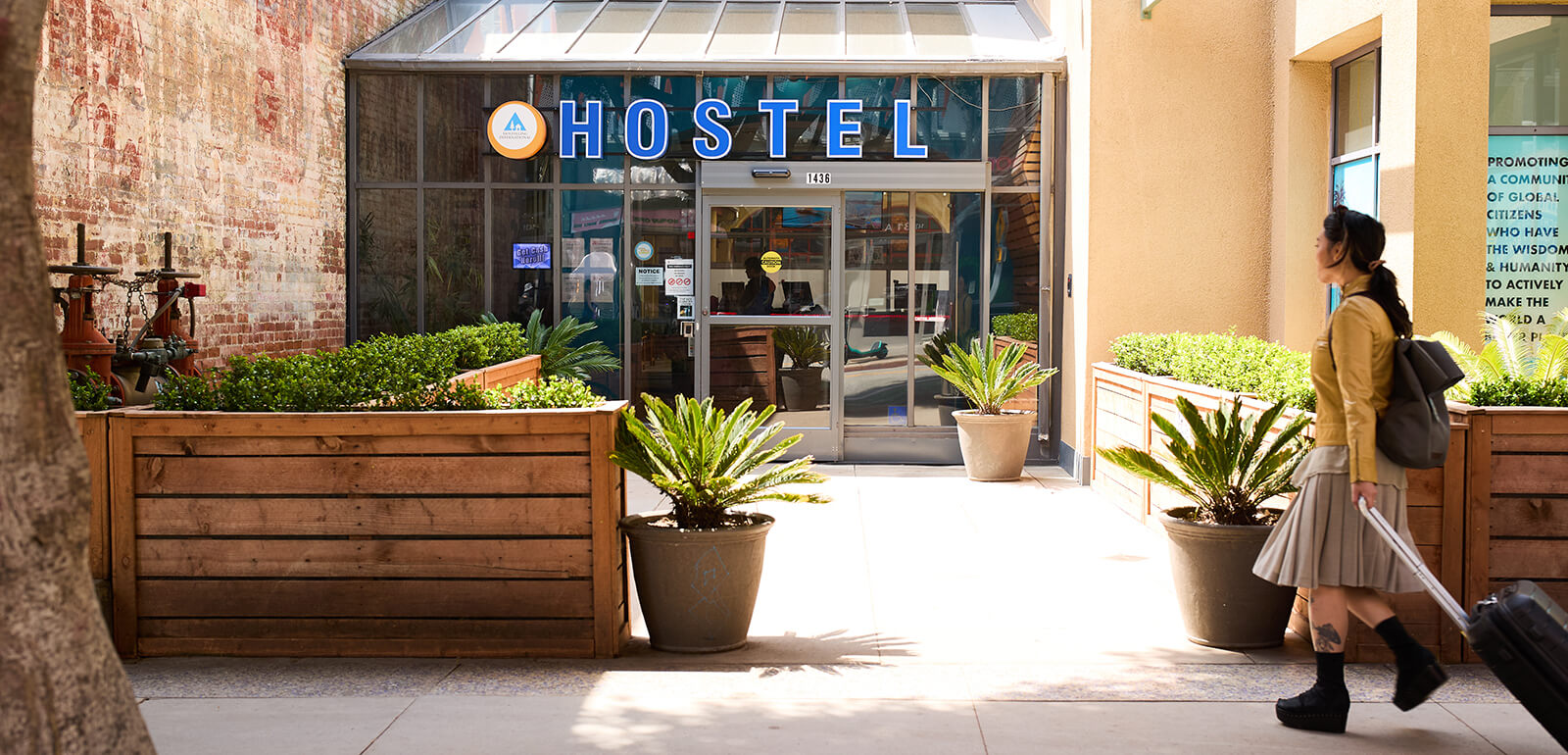 a young traveler in a cream-colored jacket, grey skirt, and black boots wheels a suitcase towards the entrance of a HI USA hostel