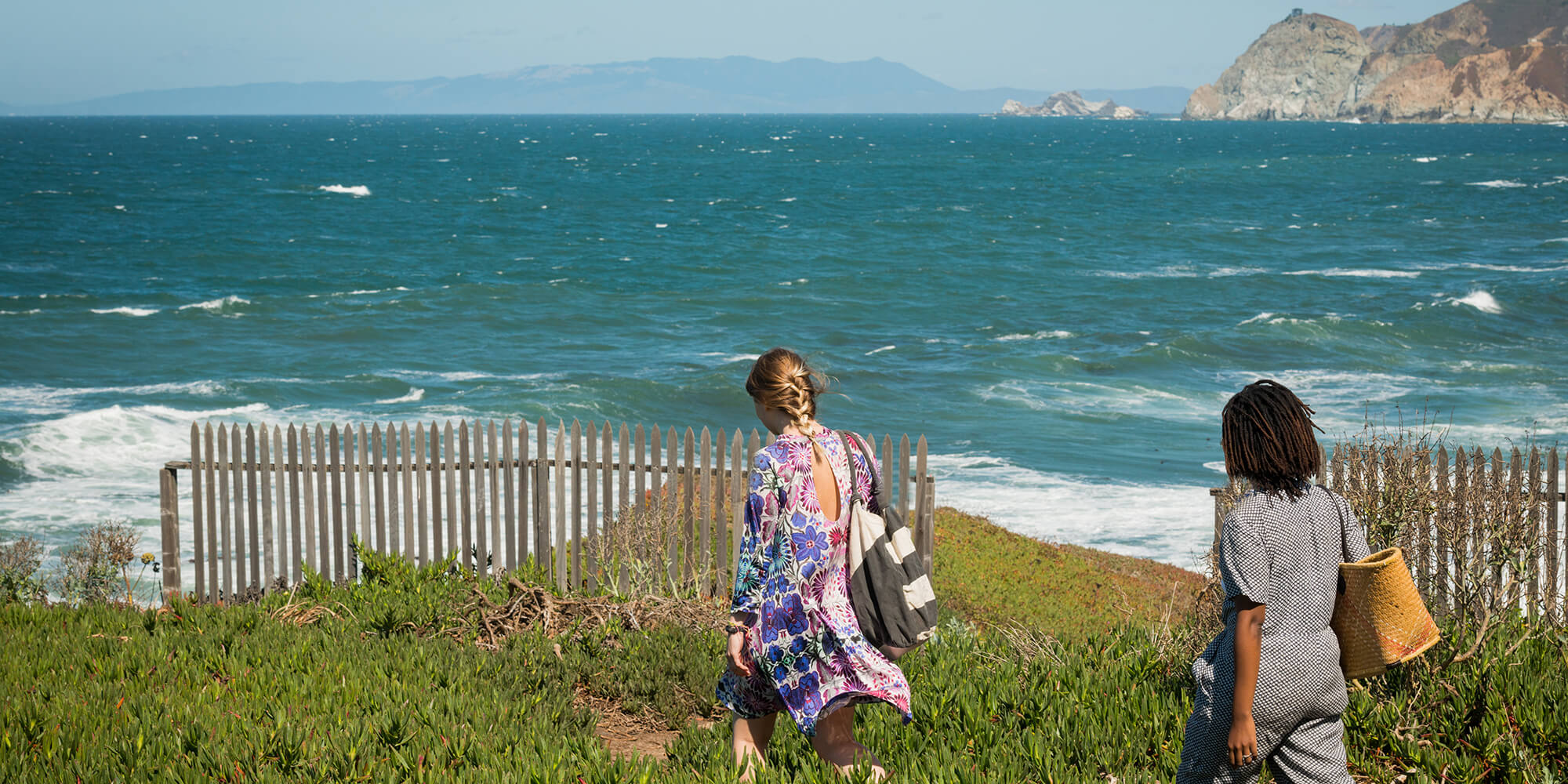 Two travelers walk down a path through a hillside covered in succulents towards the ocean