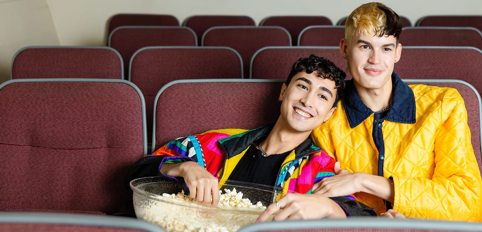 two young men in colorful jackets eat popcorn while sitting smiling in theater-style seats in a hostel movie room