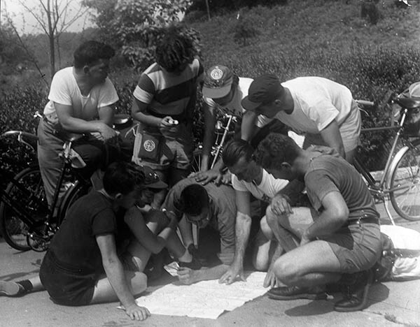 a black and white image of a group of half a dozen hostellers standing next to their bicycles and examining a trail map laid out on the ground