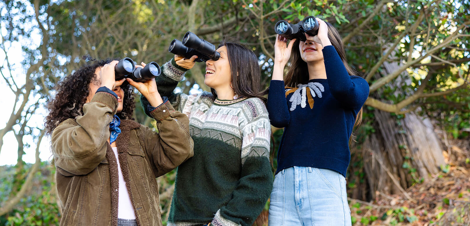 three young women hold binoculars up to their faces while standing in front of a grove of trees. Each person looks in a different direction.