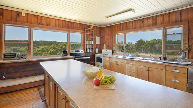 The kitchen at HI Truro hostel has a long kitchen island in the center, with wide windows overlooking the greenery and ocean outside.