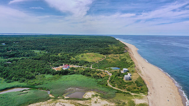 An aerial view of HI Truro Hostel sitting on a green hill overlooking a long white-sand beach running along the blue ocean.