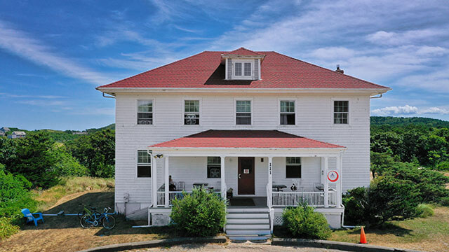The exterior of HI Truro hostel. The building is an old Coast Guard life-saving station with white exterior walls and terra cotta-colored roofing. The building is flanked by bushes and bicycles.